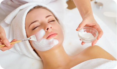 A woman lying down with her eyes closed, wearing a white towel headwrap, is receiving a facial treatment. An unseen individual applies a white mask to her face using a brush, while holding a small bowl containing the mask mixture. The setting appears to be a spa or beauty salon.