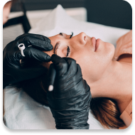 A woman lying down during a beauty procedure, with her eyes closed and a serene expression. A professional, wearing black gloves, is performing a procedure near her eyes using a specialized tool. The setting appears to be a spa or beauty salon, and the focus is on the woman's face and the hands of the professional.