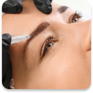 A close-up of a woman's face during an eyebrow treatment. She has well-defined eyebrows, and a practitioner wearing black gloves is applying a solution using a transparent syringe. The woman's eye is open, showcasing her eyelashes and brown iris, and she has a relaxed expression. The setting appears to be a professional beauty or cosmetic procedure environment.