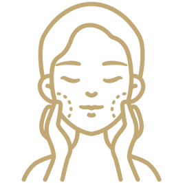 A line icon depicting the outline of a female face with closed eyes. She has flowing hair that reaches just below her ears. On her cheeks, there are dotted patterns, possibly representing freckles or skin treatments. The design is in a gold color 